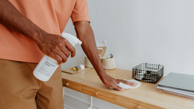 A person cleaning a wooden desk with a spray bottle and cloth, showcasing tidiness.