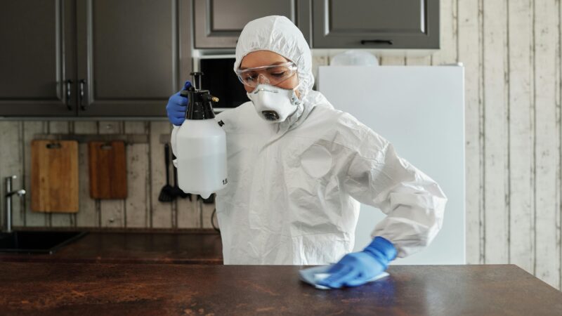 A person in protective clothing disinfects a kitchen counter indoors, focusing on safety and cleanliness.